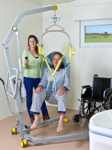 Man suspended in sling being transferred to wheelchair by attendant using Surehands Mobile Lift (model 1640).