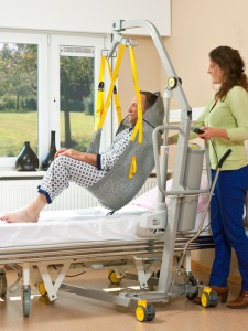 Man suspended in sling being transferred to bed by attendant using Surehands Mobile Lift (model 1630).